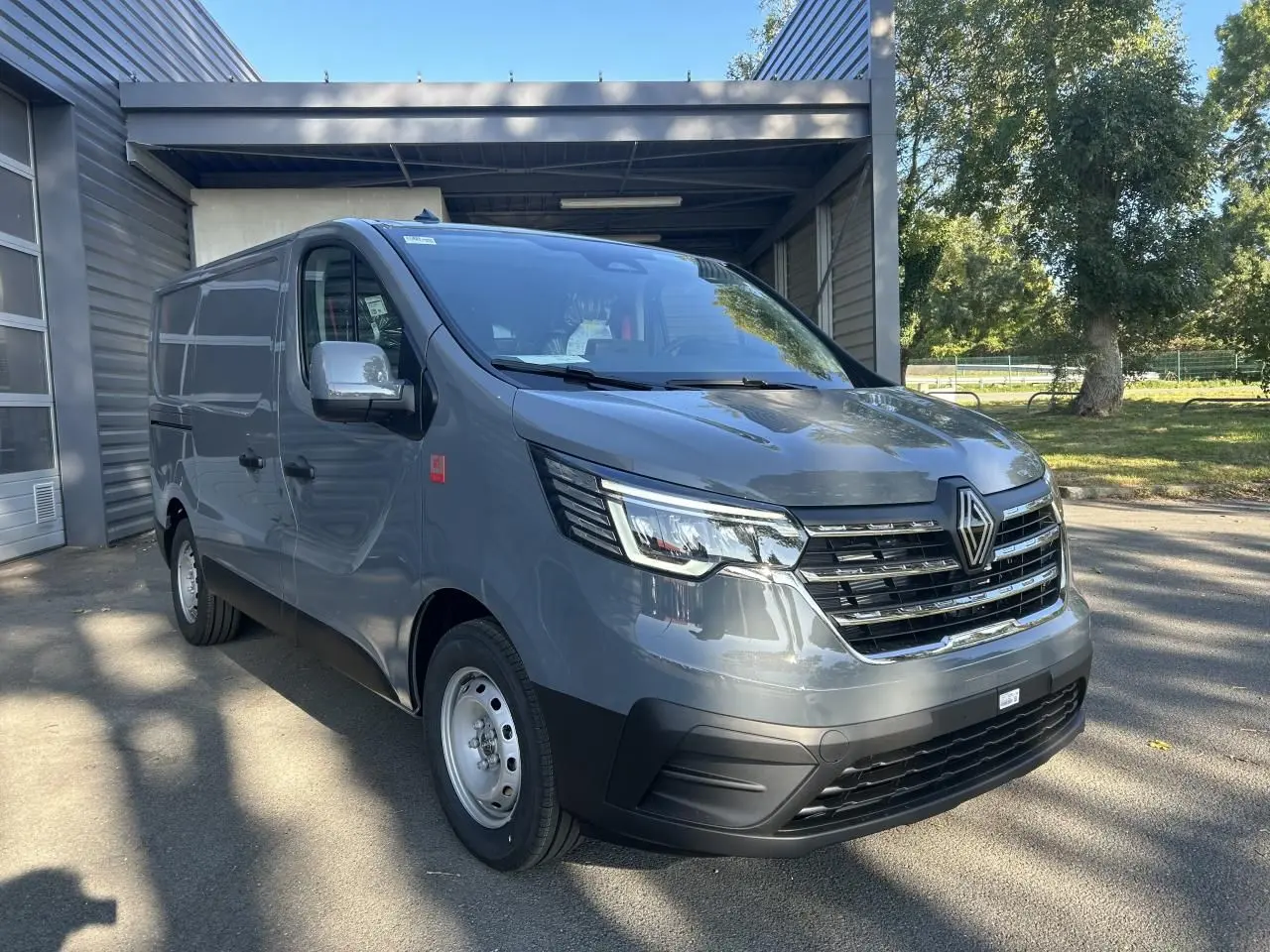 Renault Trafic Fourgon gris urbain en 3/4 avant droit, avec calandre chromée et caméra de recul visible.