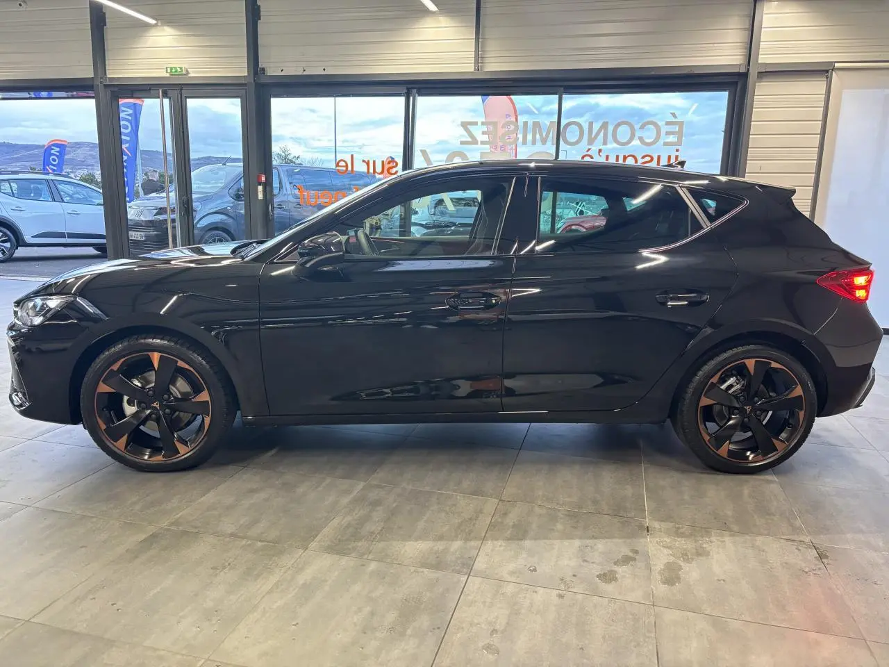 Vue de profil côté gauche d'une CUPRA Leon noir midnight avec jantes alliage 18'' cuivrées dans un showroom.
