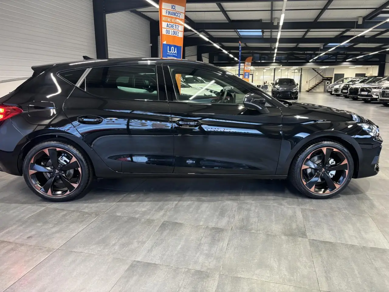 Profil droit d'une CUPRA Leon noir midnight avec jantes alliage 18'' cuivrées en showroom lumineux.