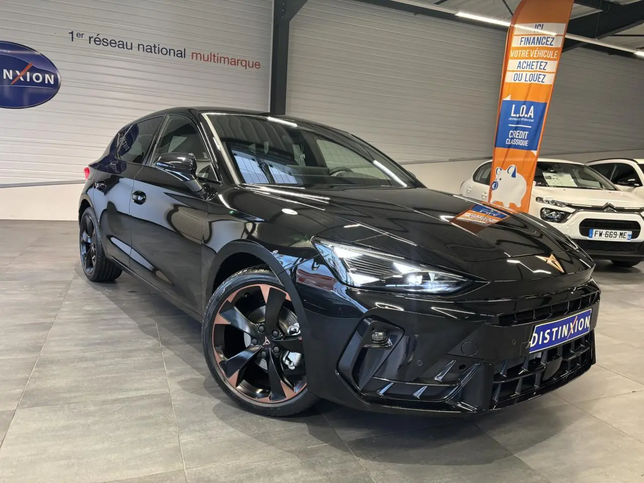 Vue 3/4 avant d'une CUPRA Leon noire midnight avec jantes alliage 18'' cuivrées et phares LED allumés en intérieur showroom.