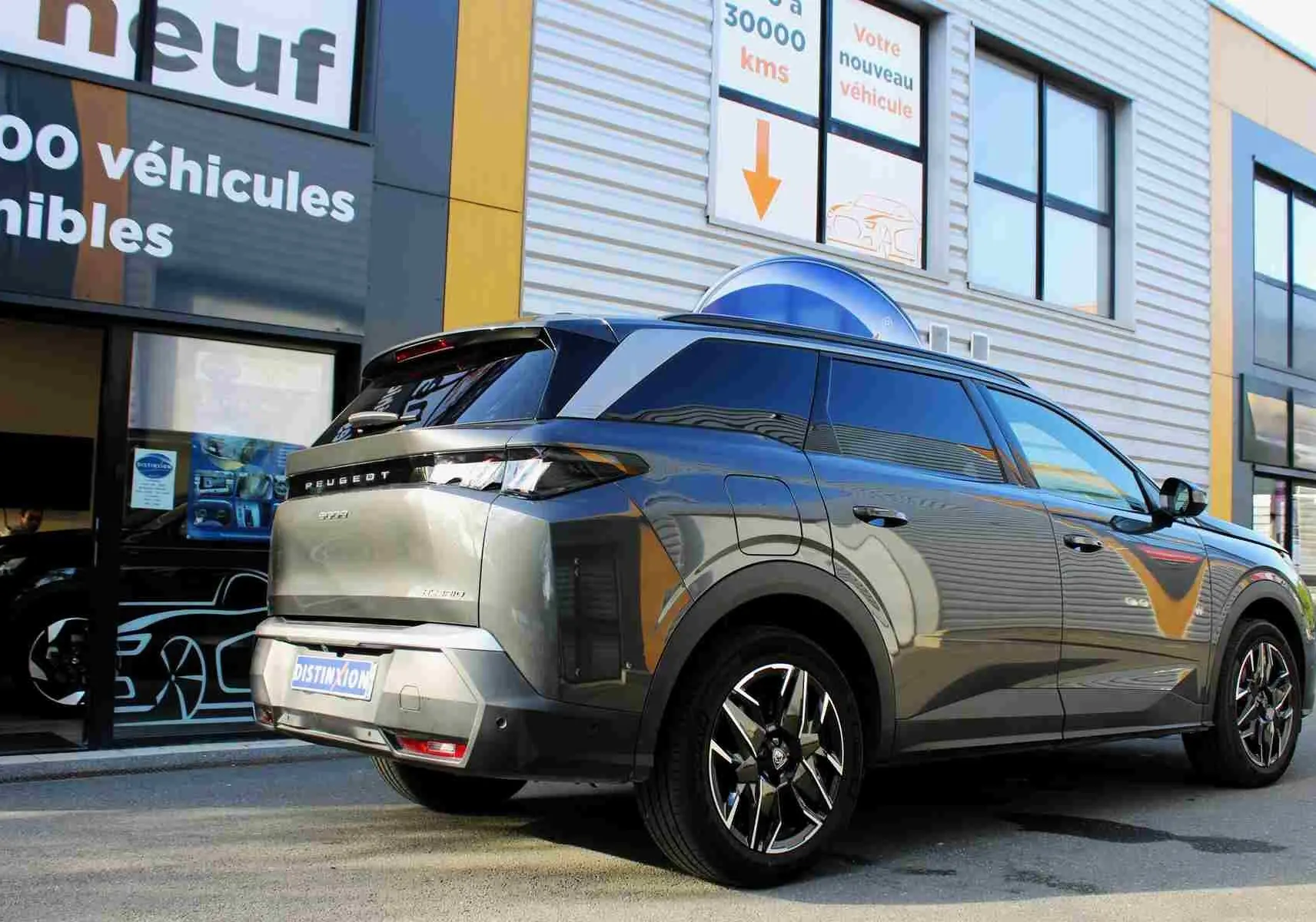 Vue 3/4 arrière droite d’un Peugeot 5008 gris titane métal avec jantes alliage et toit noir, stationné devant un showroom.
