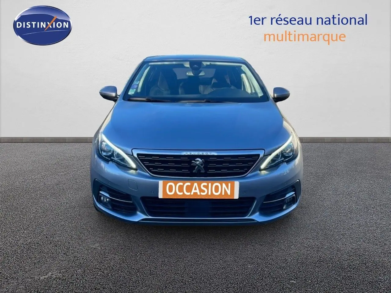 Vue frontale d'une Peugeot 308 gris Artense métal de 2019 avec phares allumés et plaque "OCCASION" visible.