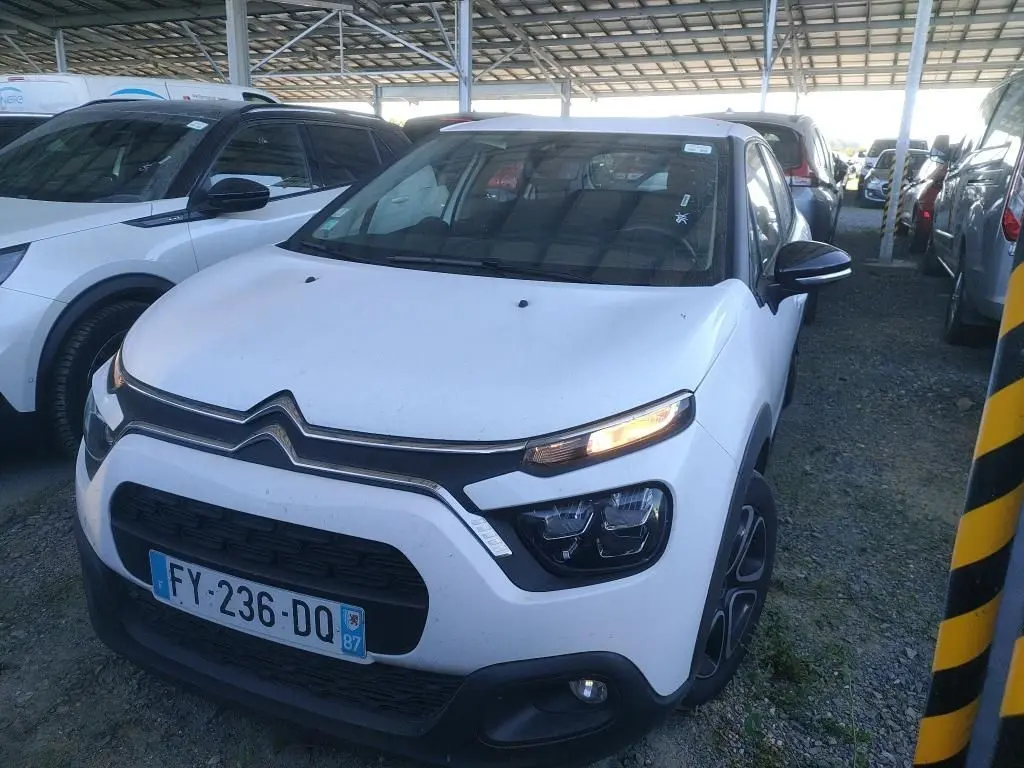 Vue 3/4 avant d'une Citroën C3 Société blanche de 2021 avec feux allumés, garée sous abri sur sol gravillonné.