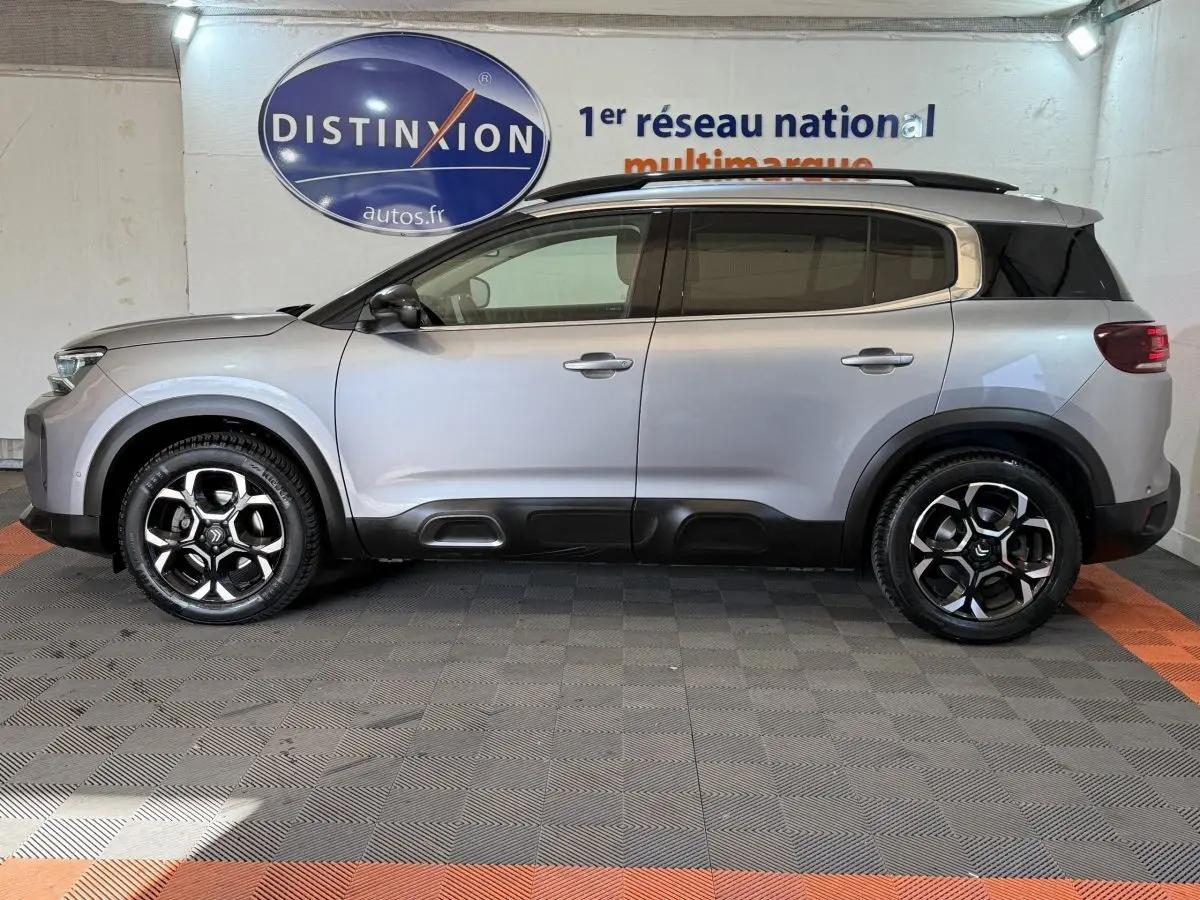Vue de profil côté gauche d’un Citroën C5 Aircross gris clair avec jantes noires et toit noir en intérieur showroom.