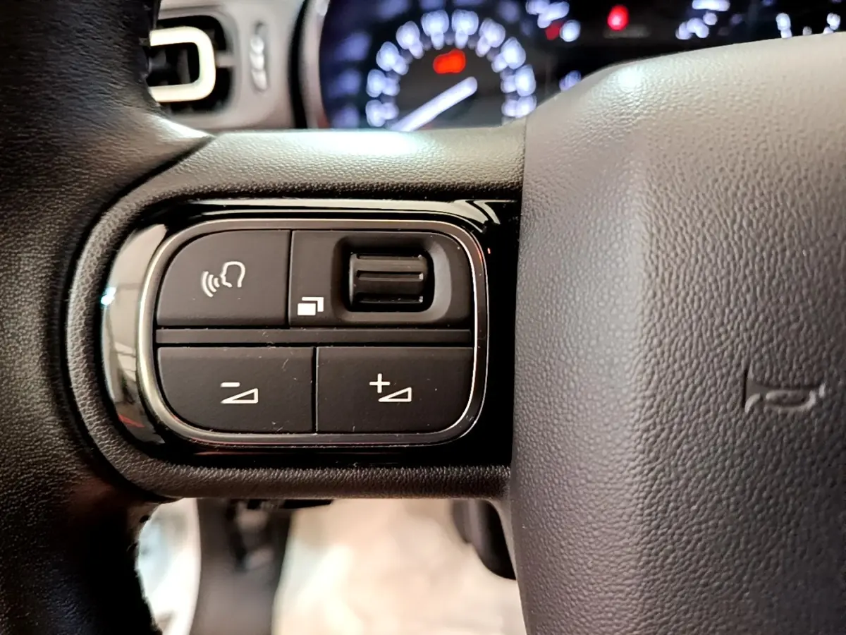 Gros plan sur les commandes vocales et volume au volant cuir noir d'une Citroën C3 blanche, tableau de bord flou en arrière-plan.
