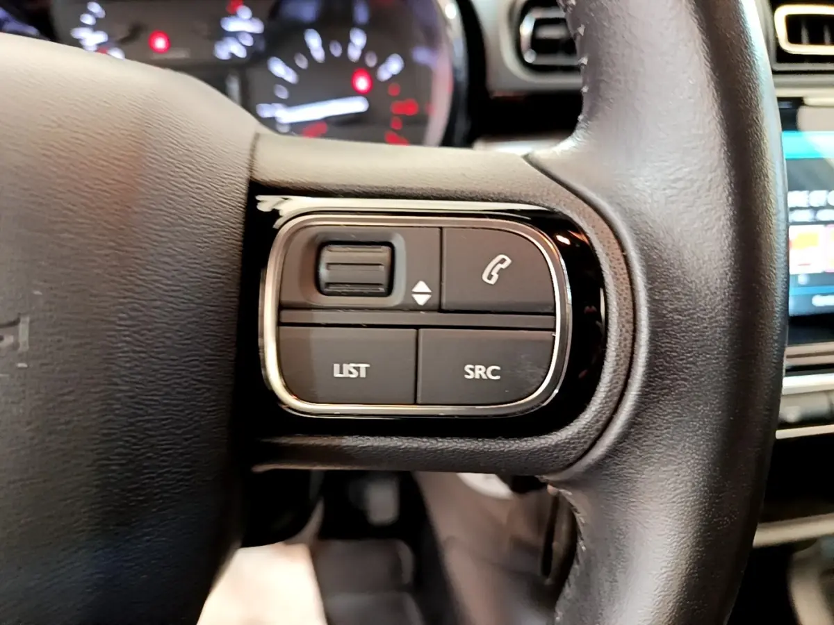 Gros plan sur les commandes gauche du volant cuir d’une Citroën C3 blanche, avec tableau de bord flou en arrière-plan.