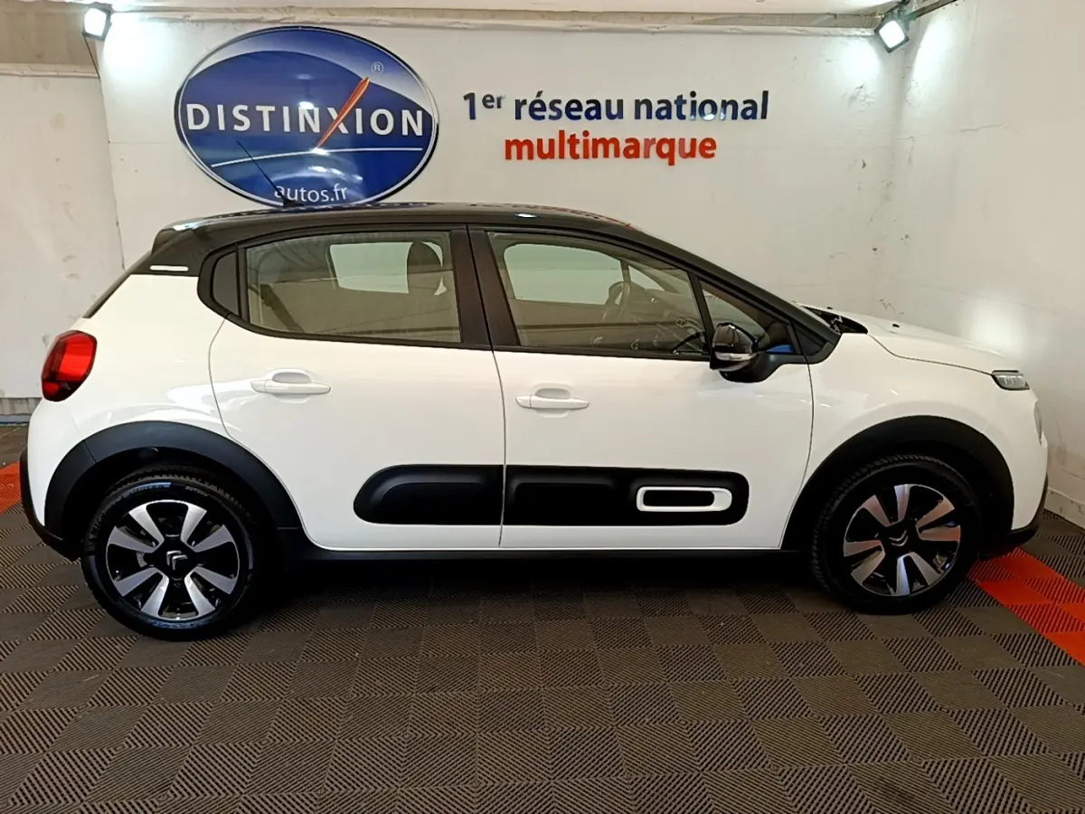 Profil latéral droit d'une Citroën C3 blanche avec toit noir et protections latérales noires, en intérieur showroom.
