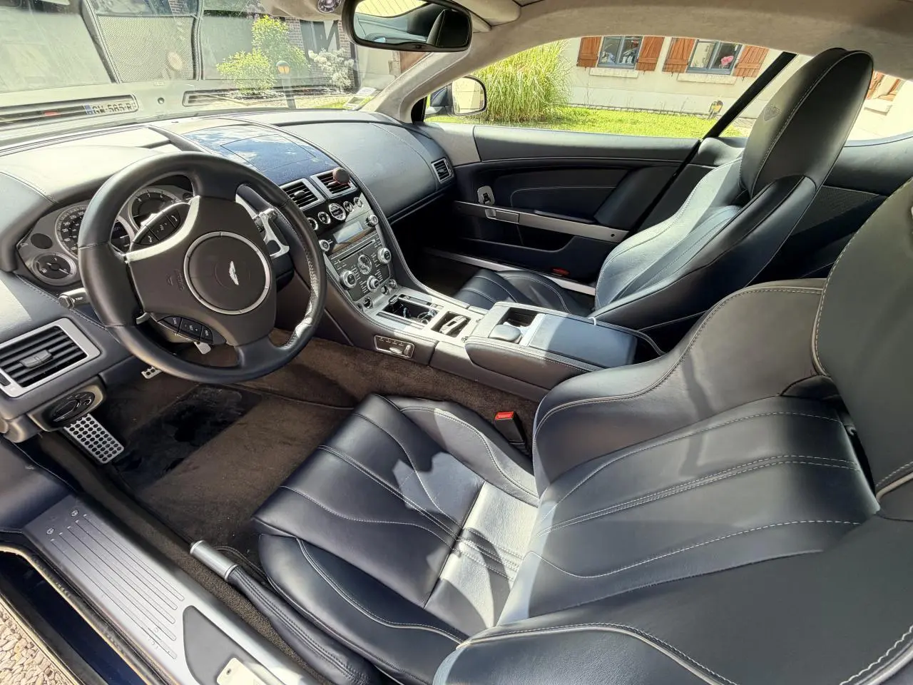 Vue intérieure côté conducteur de l'Aston Martin DB9 2016, sièges cuir noir avec coutures contrastées et tableau de bord épuré.
