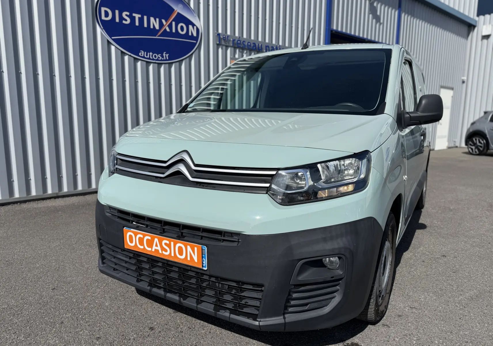 Vue 3/4 avant droit d’un Citroën Berlingo beige 2019 avec pare-chocs noir et plaque orange « OCCASION » devant un bâtiment industriel.