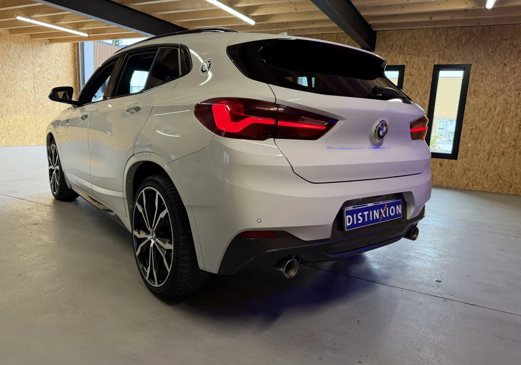 BMW X2 sDrive18d blanc vue 3/4 arrière droit en intérieur, avec jantes 19'' et doubles sorties d'échappement chromées.