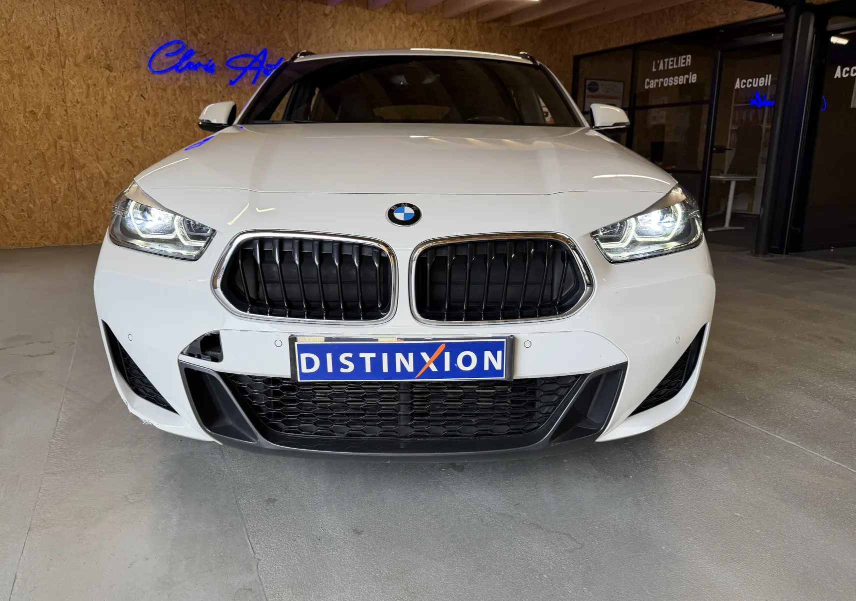 Vue frontale d'un BMW X2 blanc avec calandre noire et phares LED allumés dans un garage.