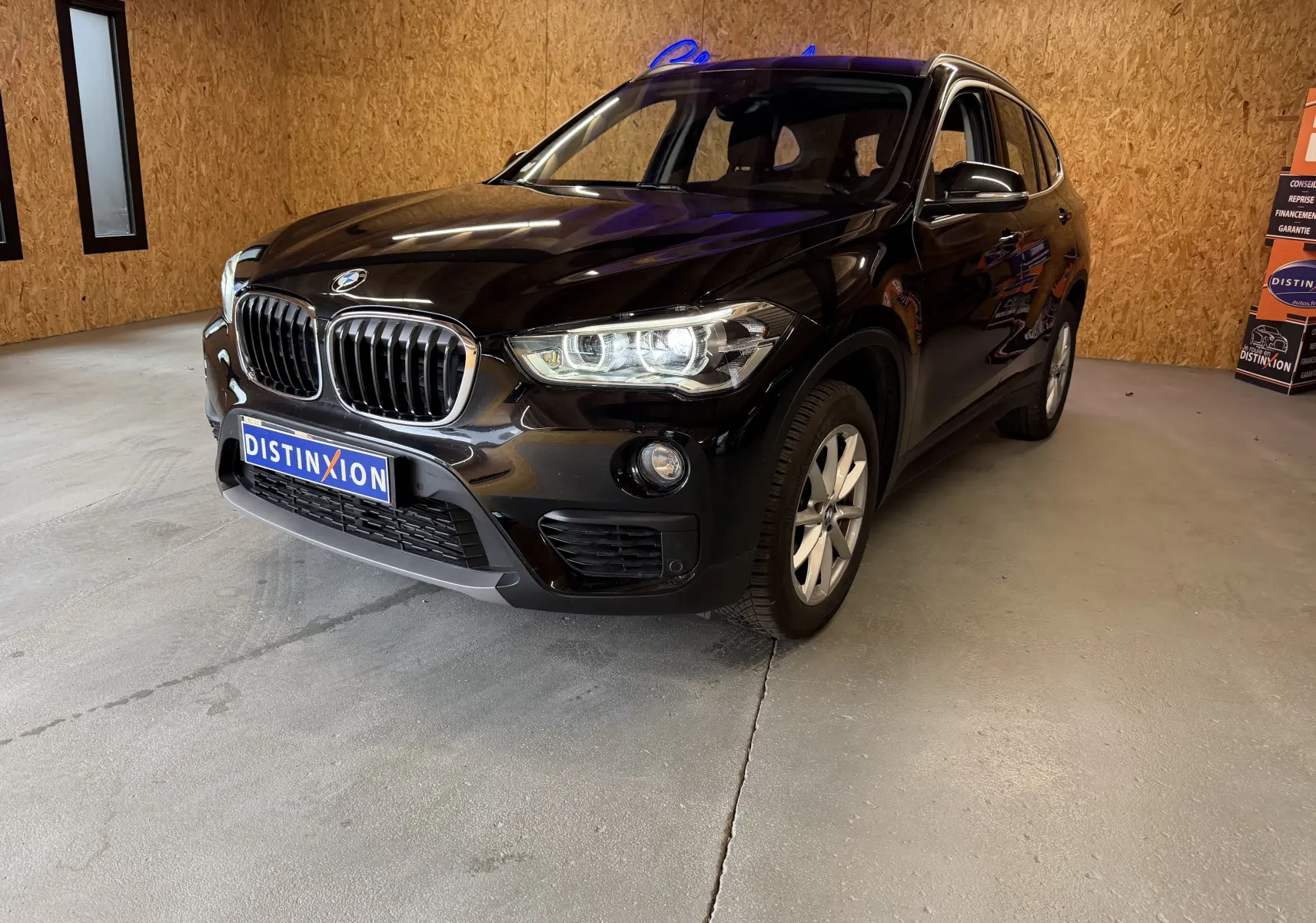 BMW X1 noir vue 3/4 avant droit dans un showroom avec calandre et phares allumés visibles.