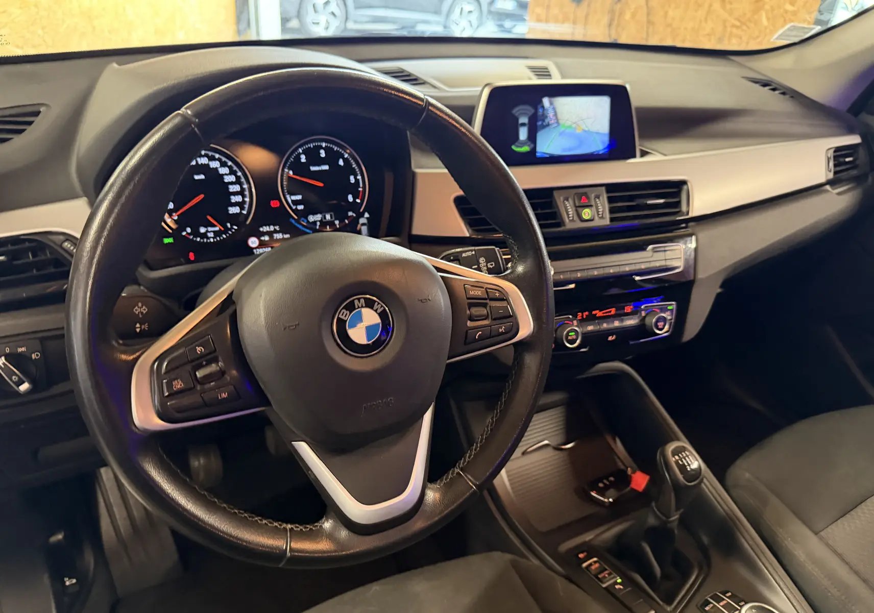 Vue intérieure centrée sur le volant et tableau de bord d'une BMW X1 noire avec boîte manuelle et écran multimédia allumé.