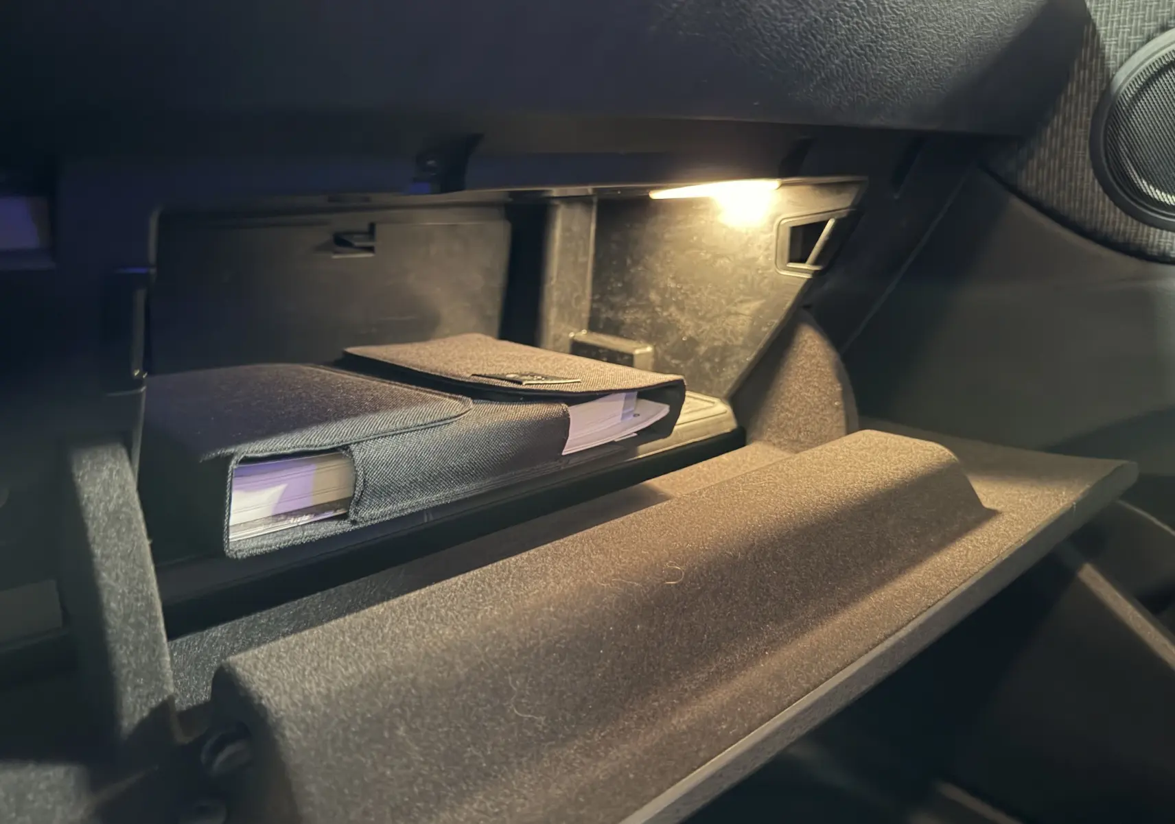 Gros plan sur la boîte à gants ouverte du BMW X1 noir 2019, avec un manuel dans une pochette textile et un éclairage intérieur.