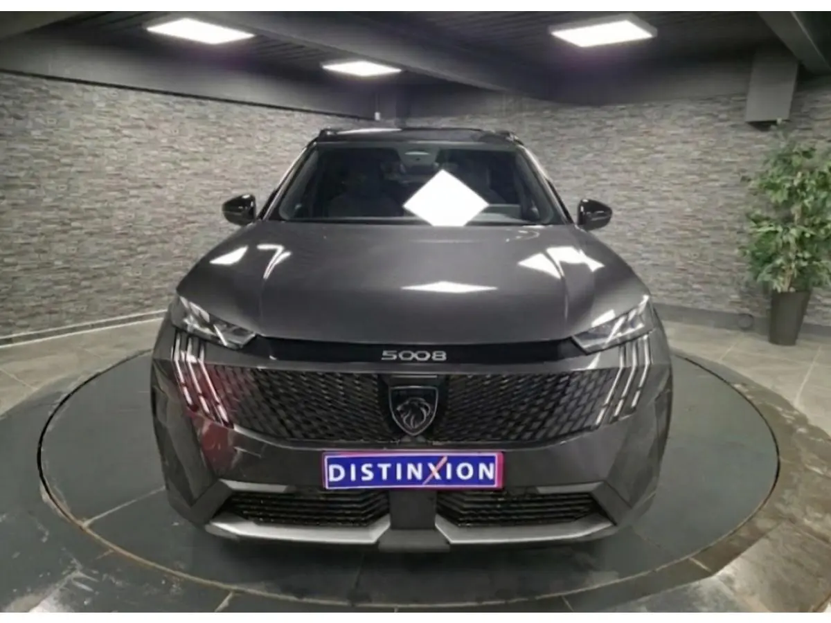 Vue avant d'un Peugeot 5008 gris métallisé 2025 avec calandre noire et feux LED à trois griffes, en intérieur.