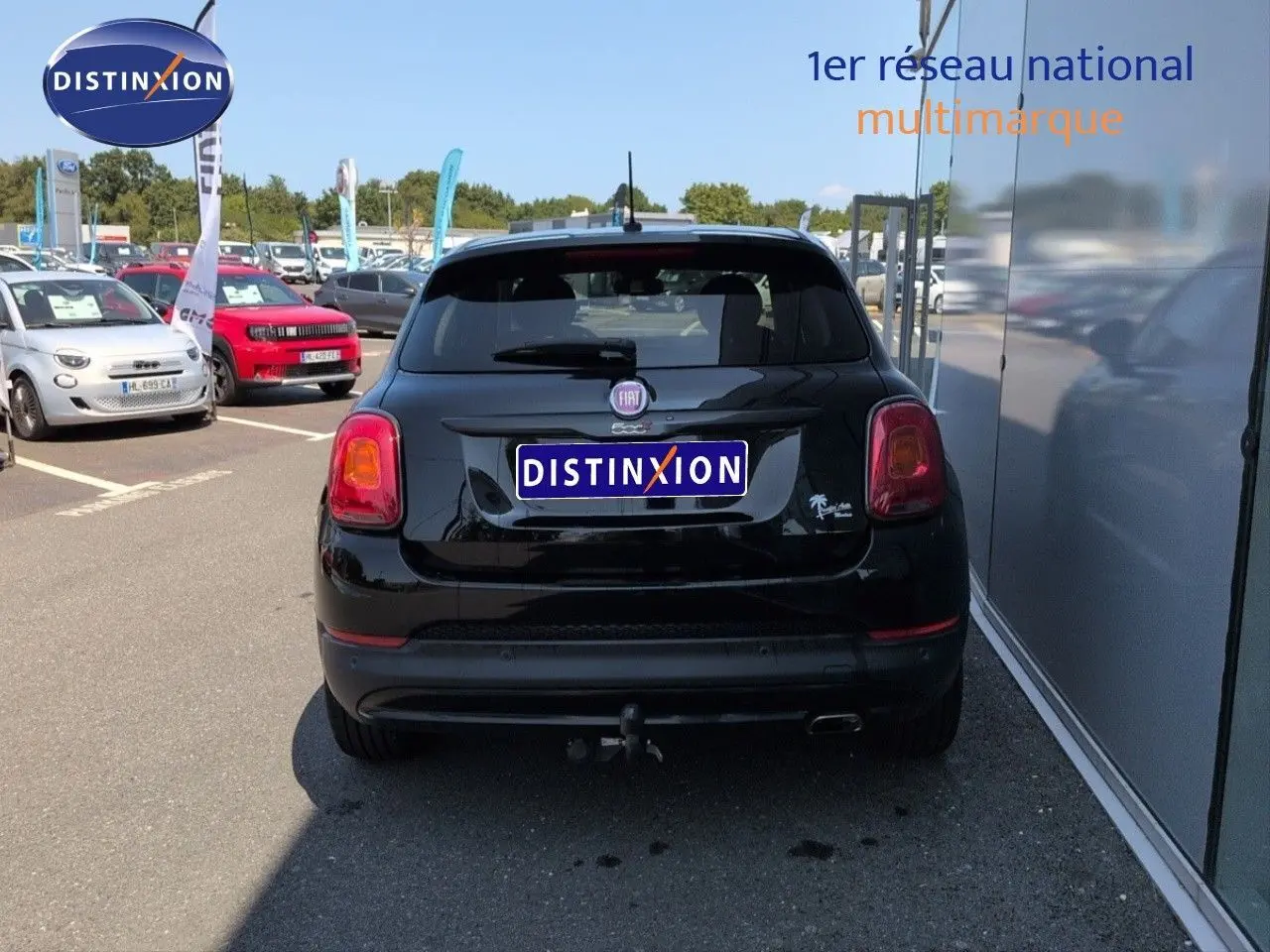 Vue arrière d'une Fiat 500X noire Cinéma pastel avec attelage, stationnée près d'un bâtiment vitré.