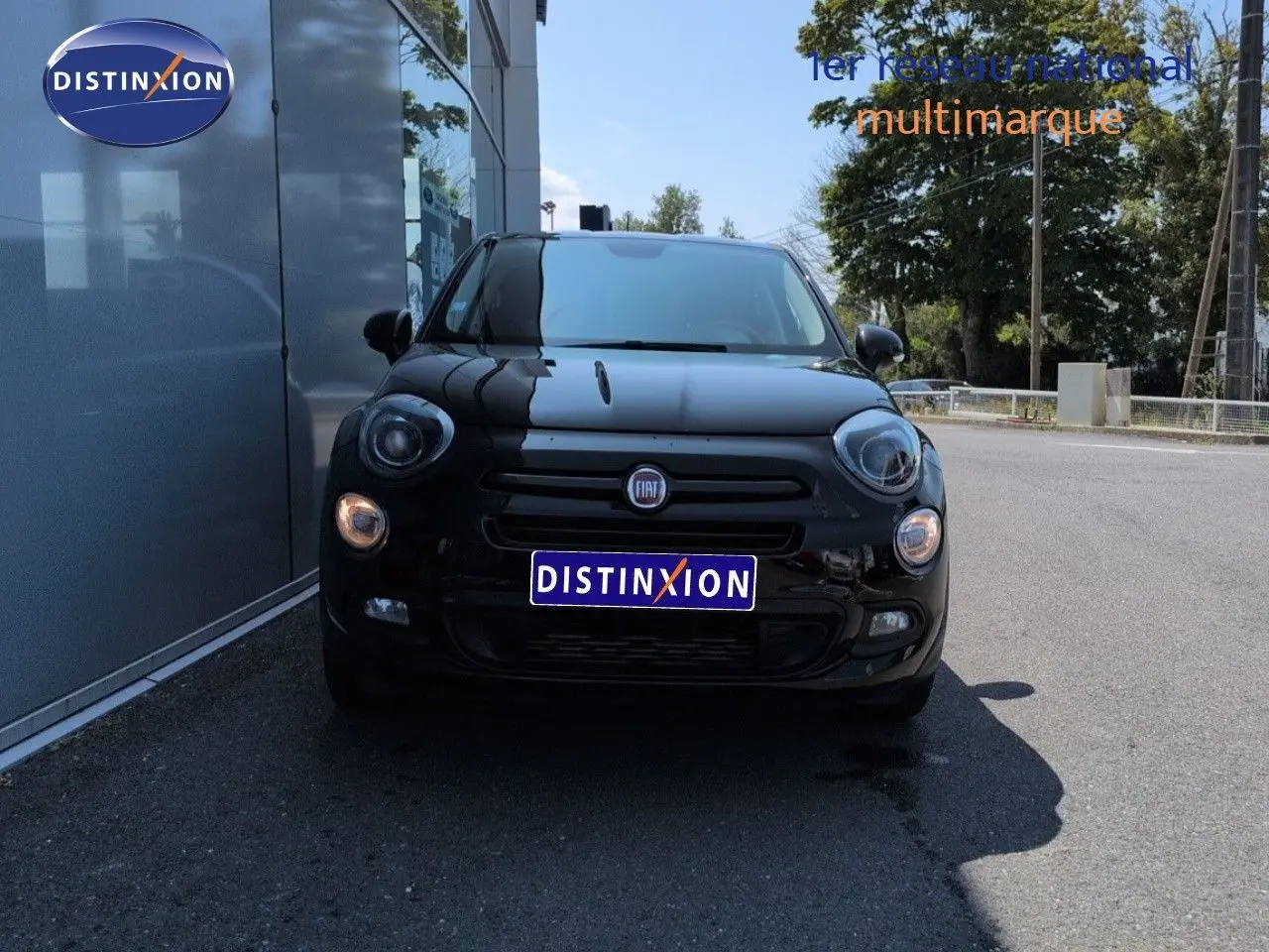 Vue de face d'une FIAT 500X noire Cinéma pastel extra-série, avec phares allumés, stationnée devant un bâtiment.