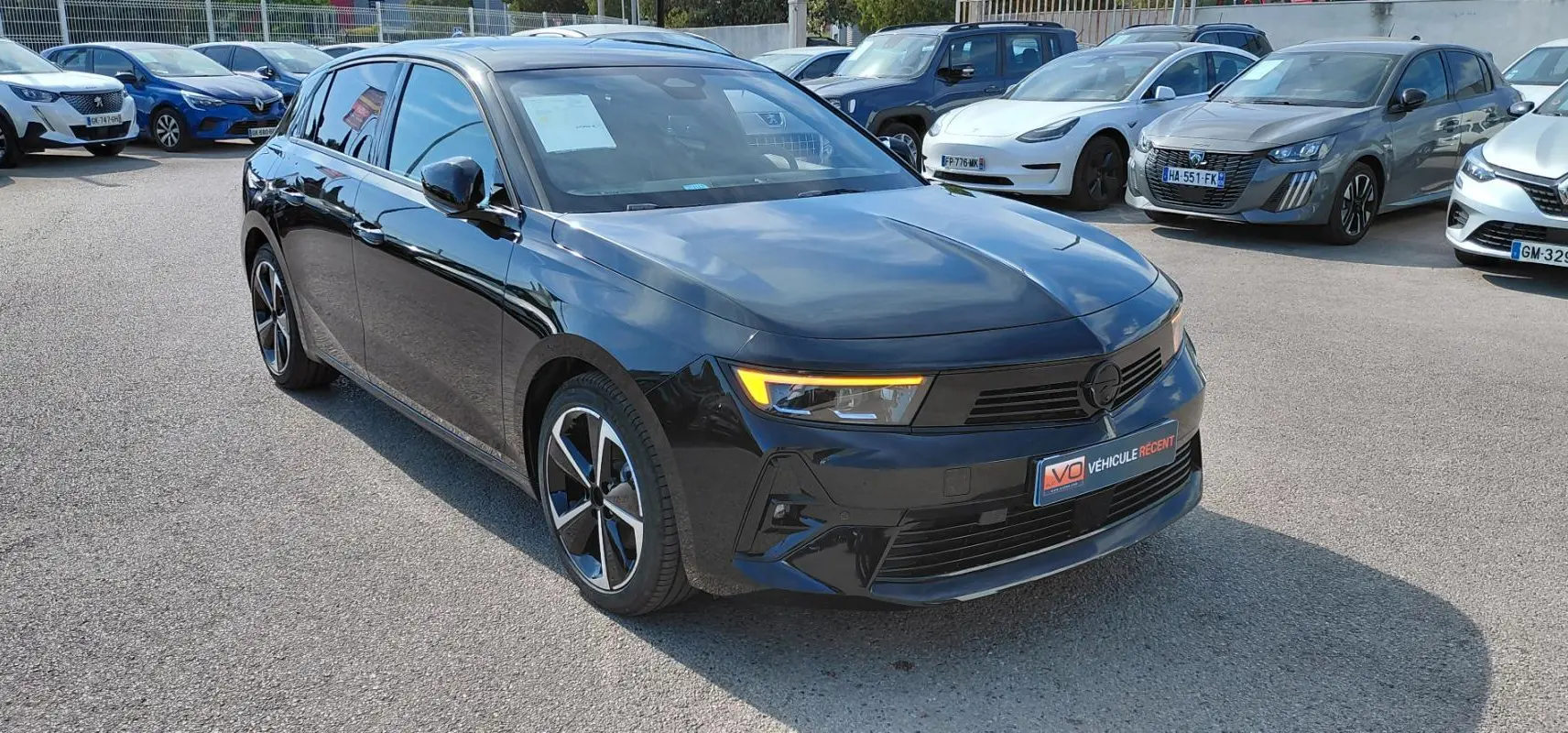 Opel Astra noire 2025 vue 3/4 avant droit avec feux diurnes allumés sur parking extérieur.