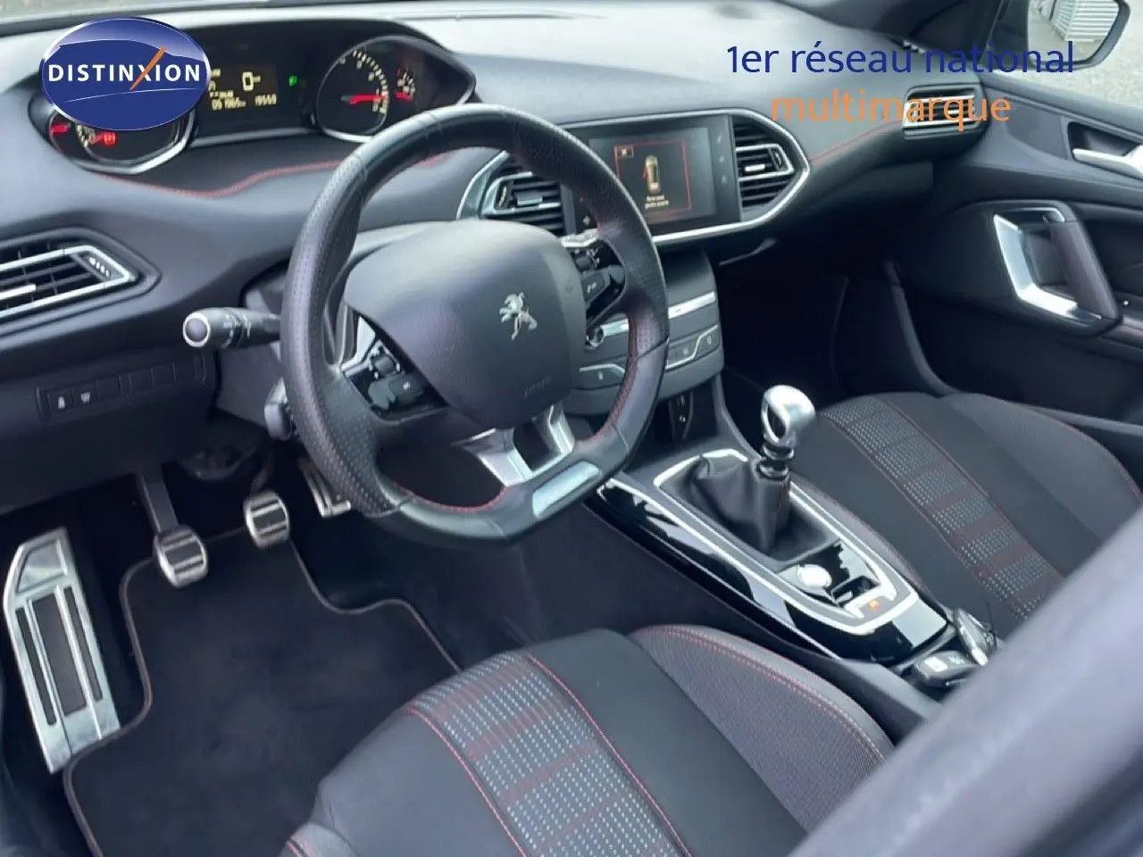 Vue intérieure côté conducteur de la Peugeot 308 gris Artense de 2017 avec volant cuir, boîte manuelle et sièges tissu noir à surpiqûres rouges.