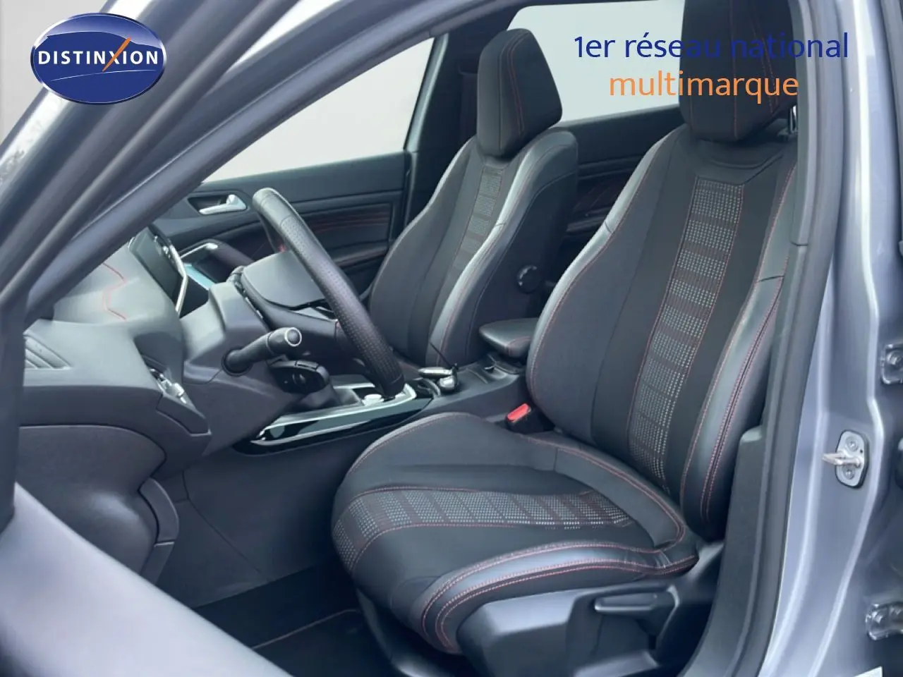 Vue intérieure côté conducteur de la Peugeot 308 gris Artense, sièges sport noirs avec surpiqûres rouges visibles.
