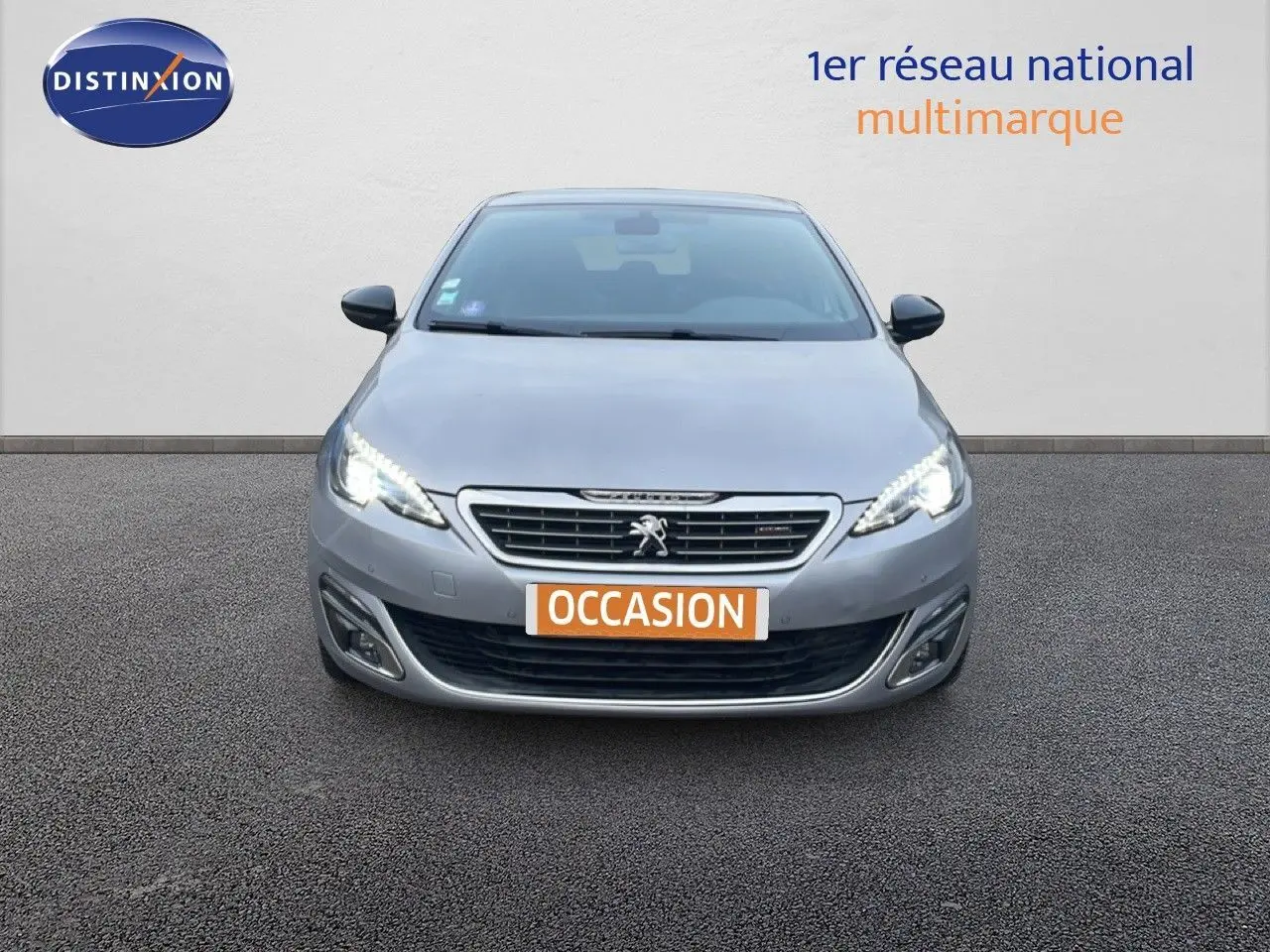 Vue de face d'une Peugeot 308 gris Artense avec phares LED allumés et plaque "OCCASION" visible.