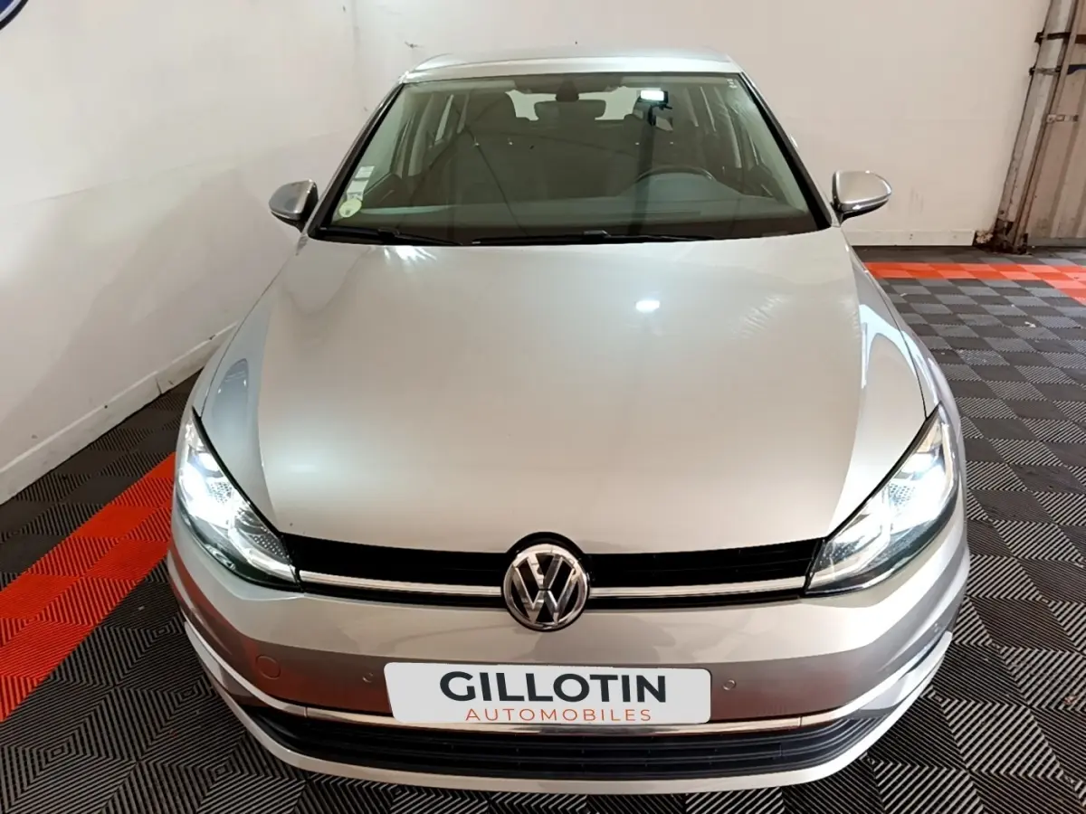 Vue avant d'une Volkswagen Golf gris clair avec phares allumés dans un garage au sol damier noir et rouge.