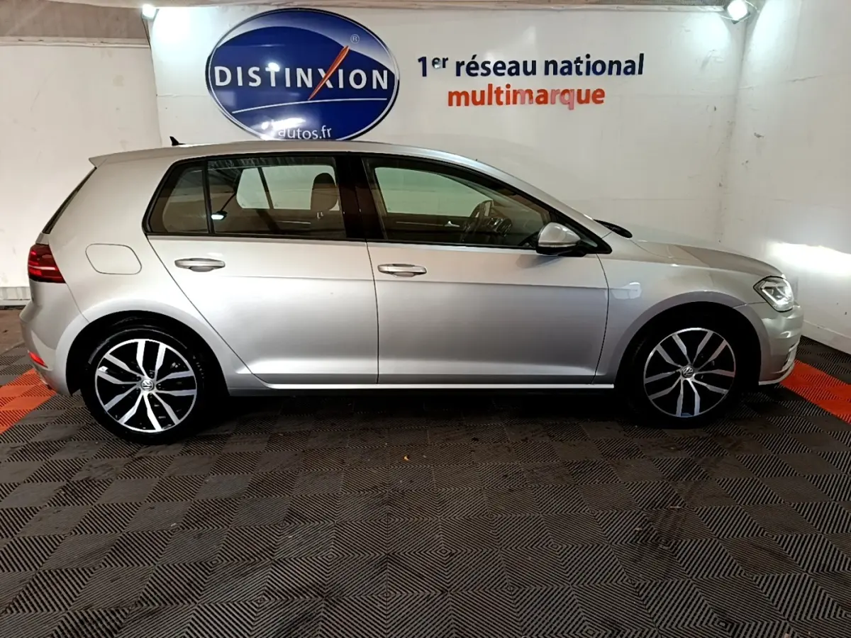 Volkswagen Golf gris clair vue de profil côté gauche, avec jantes alliage et logo Distinxion en arrière-plan.