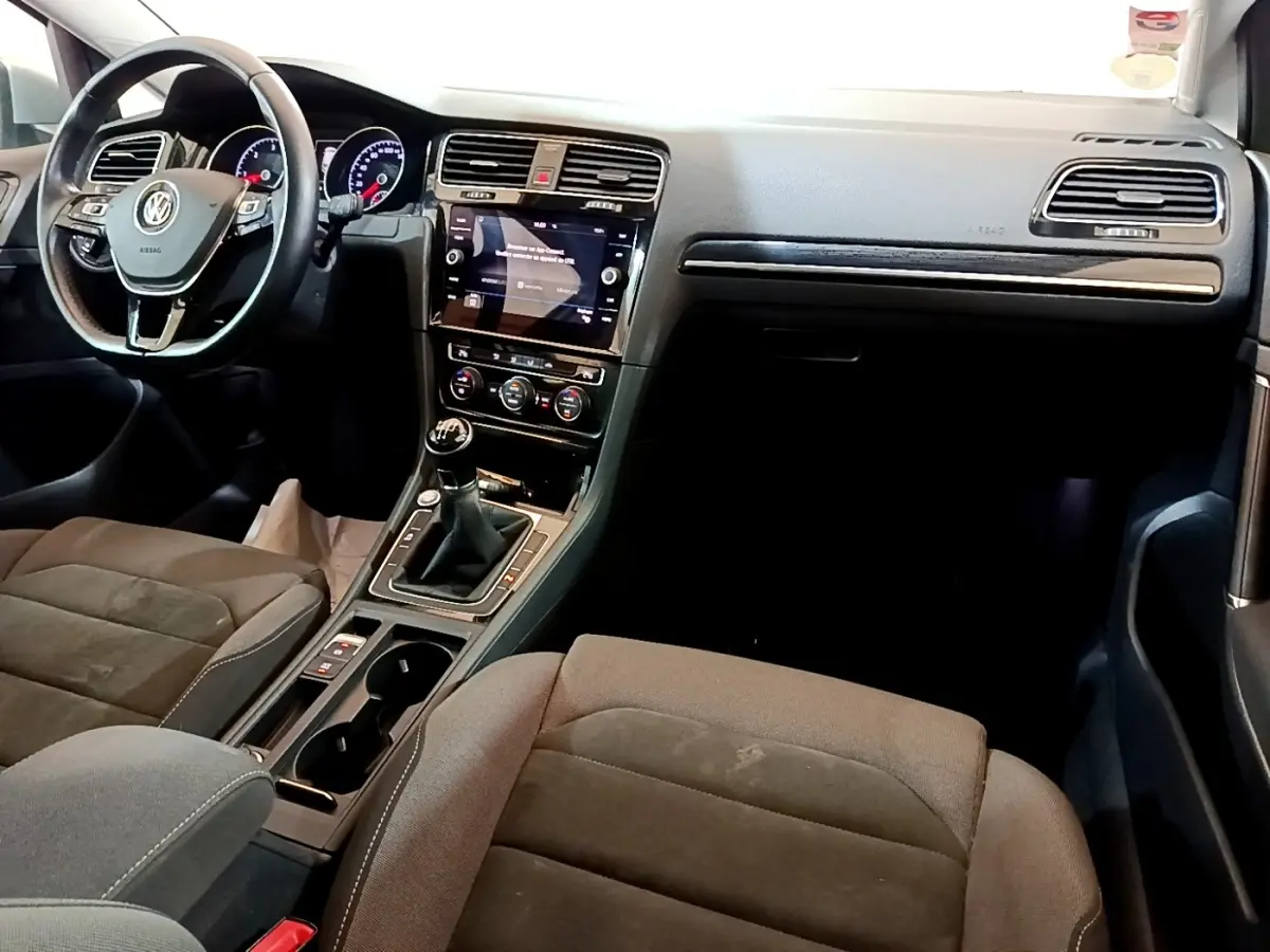 Intérieur de Volkswagen Golf gris clair, vue de face côté conducteur, volant cuir, écran tactile et boîte manuelle visible.