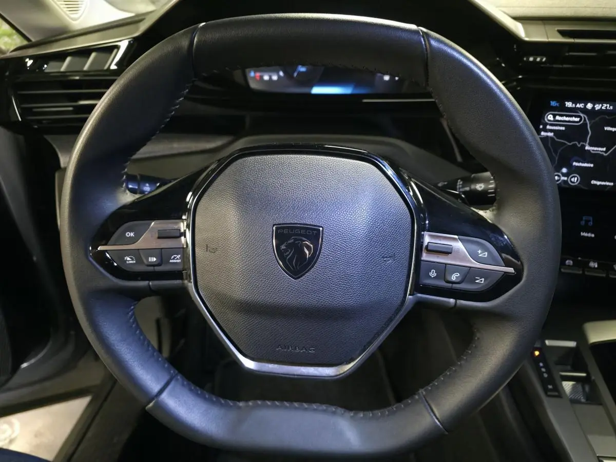 Vue rapprochée du volant cuir noir avec logo Peugeot et commandes intégrées, tableau de bord numérique en arrière-plan.