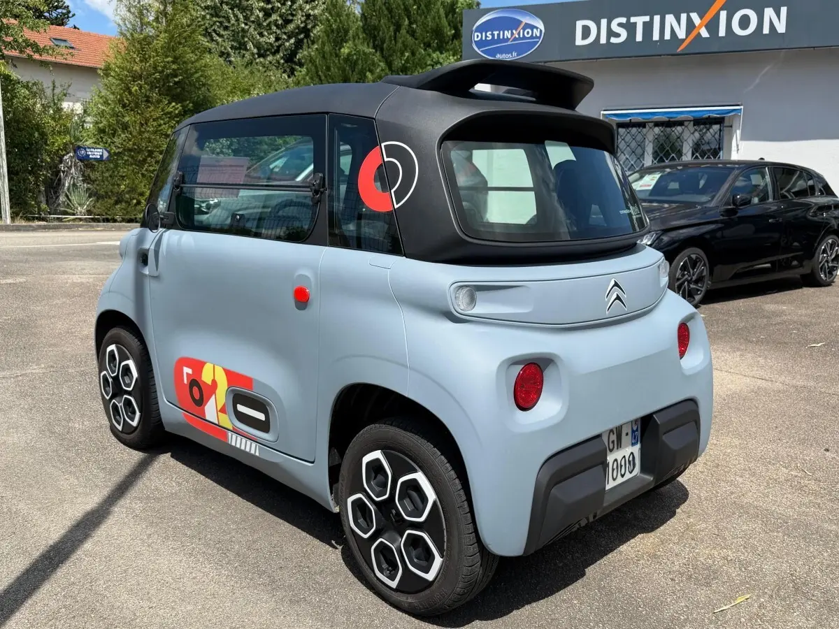 Vue 3/4 arrière droite de la Citroën Ami 2024 gris clair avec toit noir et stickers colorés sur porte et montant arrière.