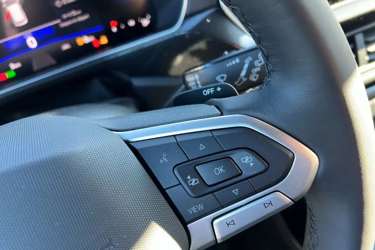 Gros plan sur les commandes multifonctions du volant cuir du Volkswagen T-Cross blanc pur, tableau de bord numérique en arrière-plan.