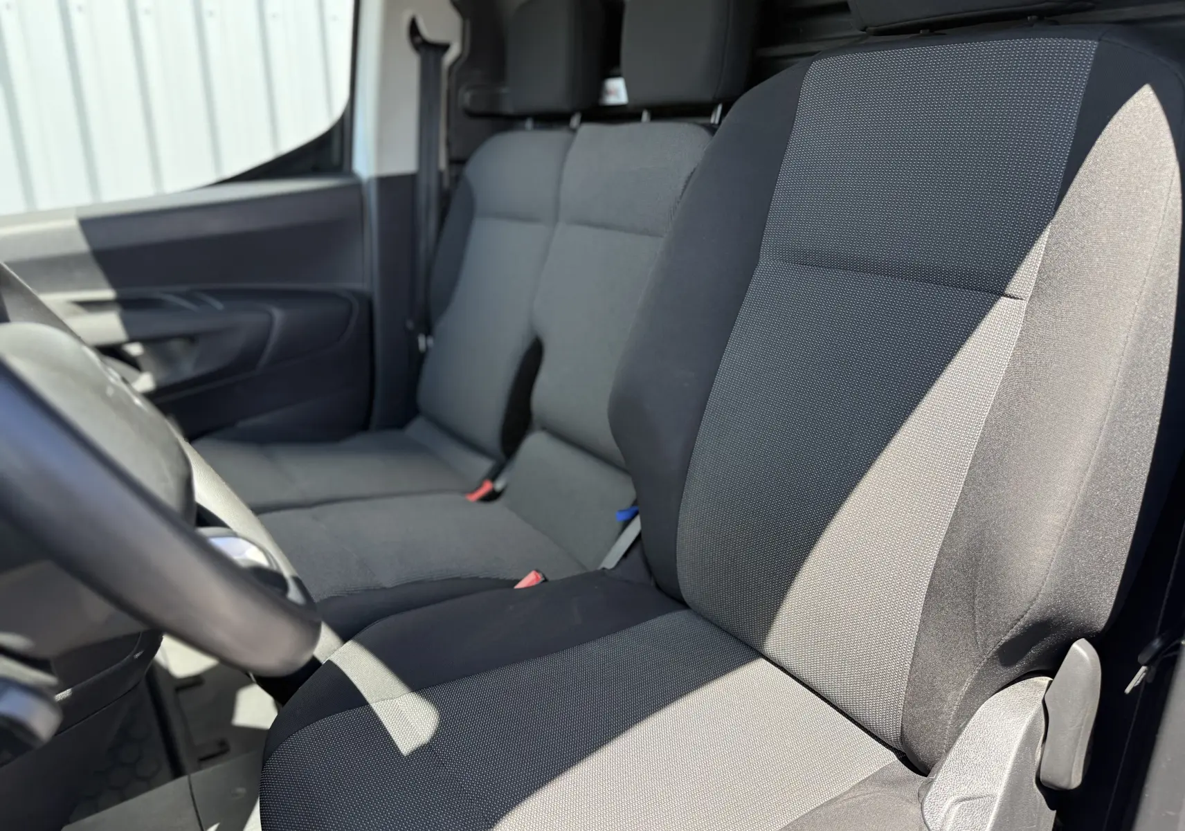 Vue intérieure côté conducteur du Citroën Berlingo XL 2022, sièges tissu gris noir avec accoudoirs et ceintures visibles.
