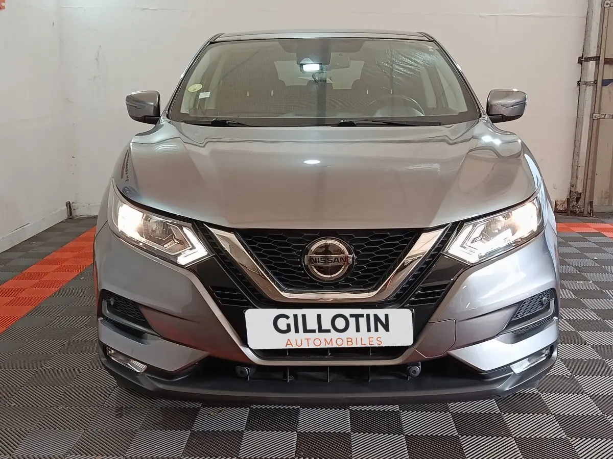 Vue avant d'un Nissan Qashqai gris clair 2019 avec phares allumés et calandre chromée dans un garage.