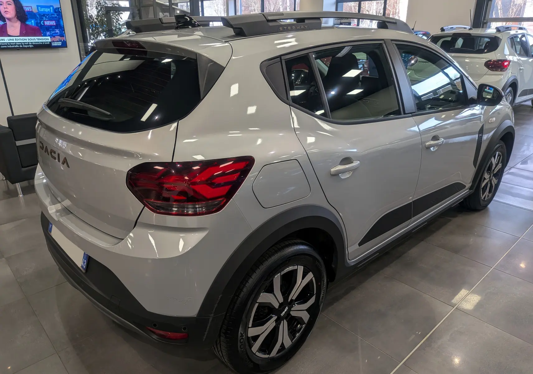 Vue 3/4 arrière droite d'une Dacia Sandero Stepway gris Moonstone en showroom, avec jantes biton et barres de toit.