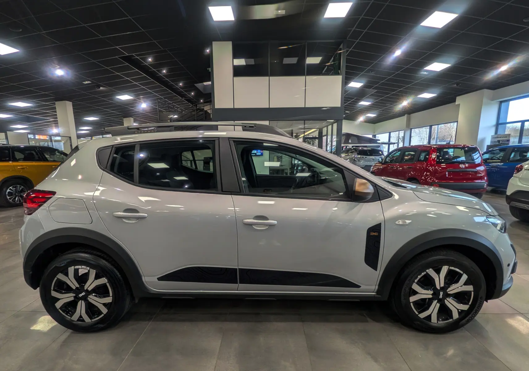 Vue de profil côté gauche d'une Dacia Sandero Stepway gris Moonstone en showroom, avec jantes bi-ton et protections noires.