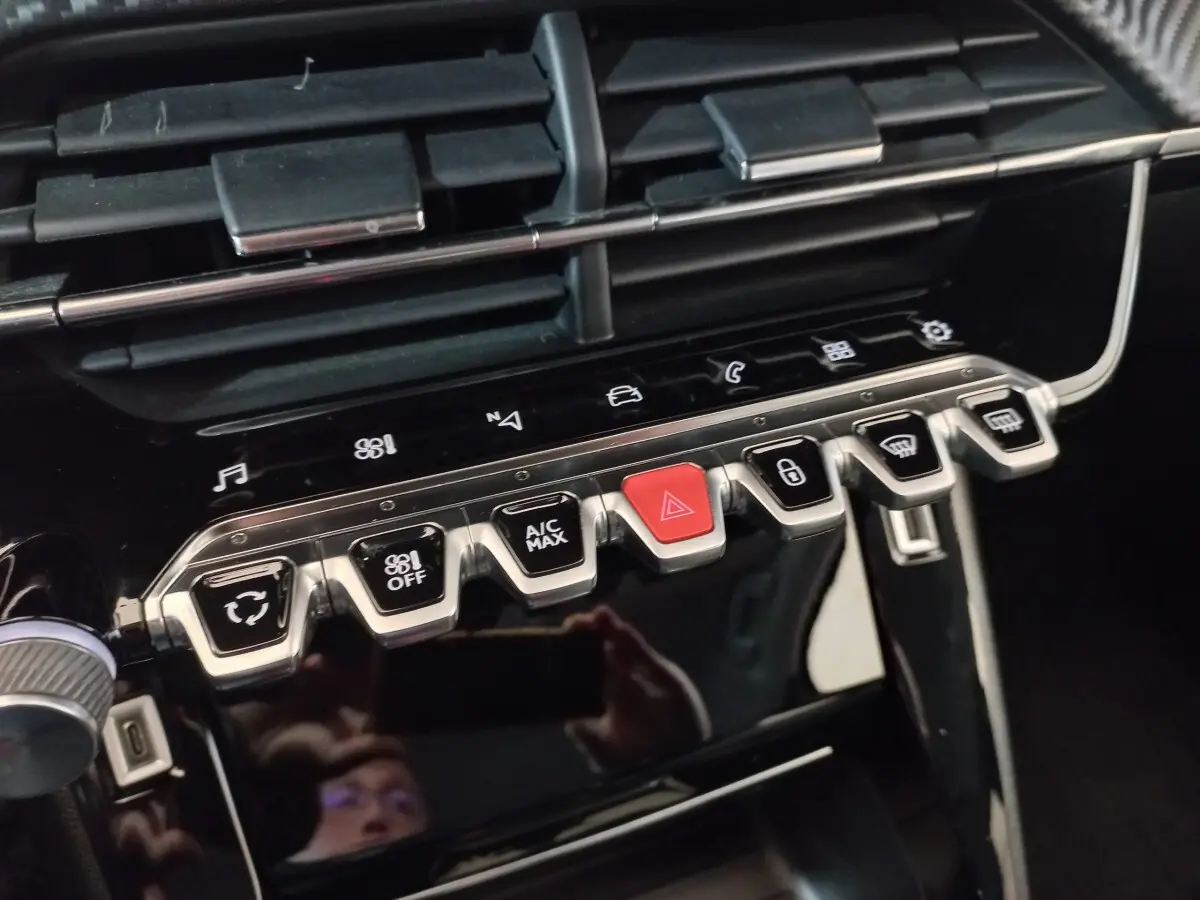 Gros plan sur la rangée de boutons chromés et noirs du tableau de bord du Peugeot 2008 orange, avec bouton rouge d’alerte au centre.