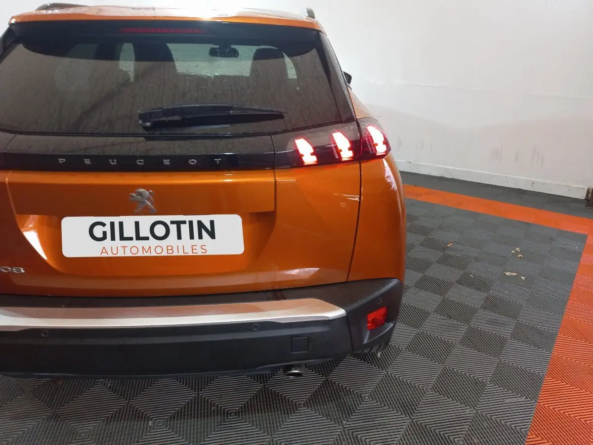 Vue arrière d'un Peugeot 2008 orange avec feux arrière allumés dans un garage au sol gris et orange.
