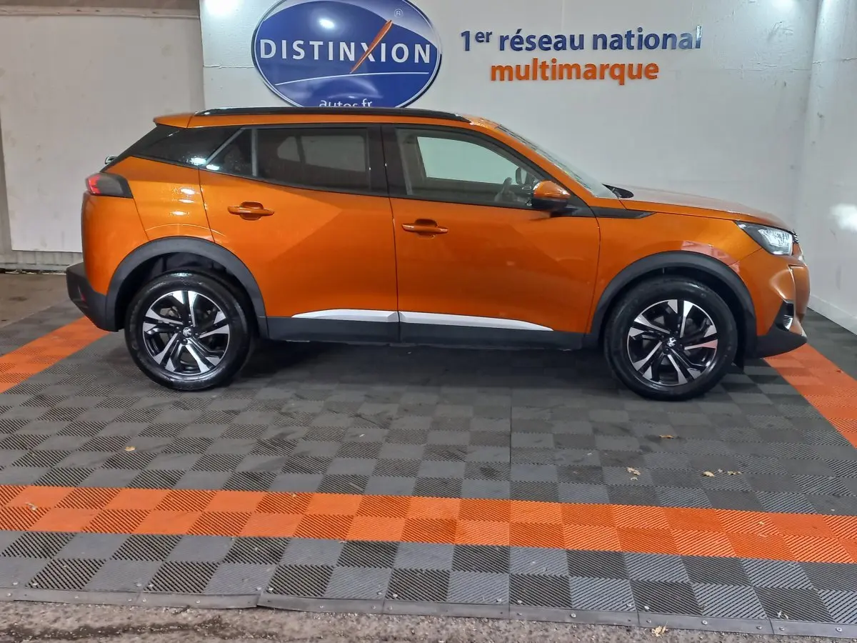 Profil droit d'un Peugeot 2008 orange 2021 avec jantes noires et toit noir dans un showroom multimarque.