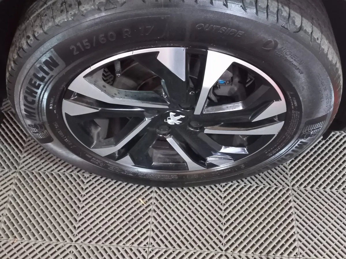 Gros plan sur la jante alliage noire et argentée avec logo Peugeot au centre, pneu Michelin visible sur sol carrelé.