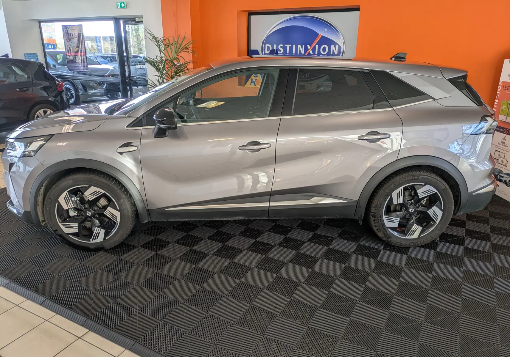Profil droit du Renault Symbioz gris cassiopée métal 2025 en hybride essence, avec jantes noires et argentées en intérieur showroom.