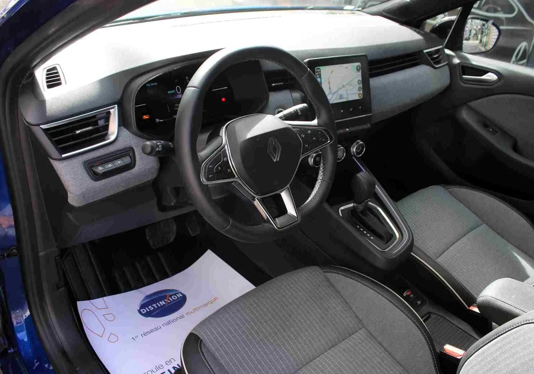 Vue intérieure côté conducteur de la Renault Clio bleu iron metal, volant multifonction et écran tactile central visible.