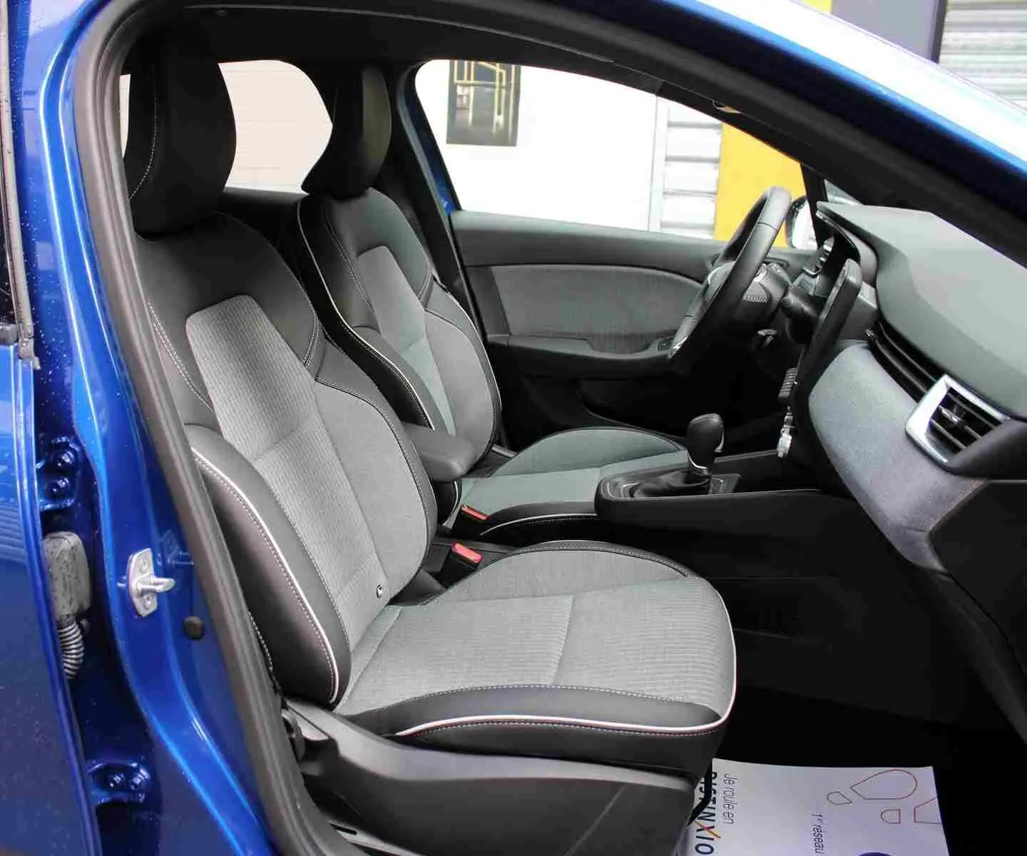 Vue intérieure côté gauche de la Renault Clio 2025 bleu iron metal, montrant les sièges avant gris et noir avec surpiqûres blanches.