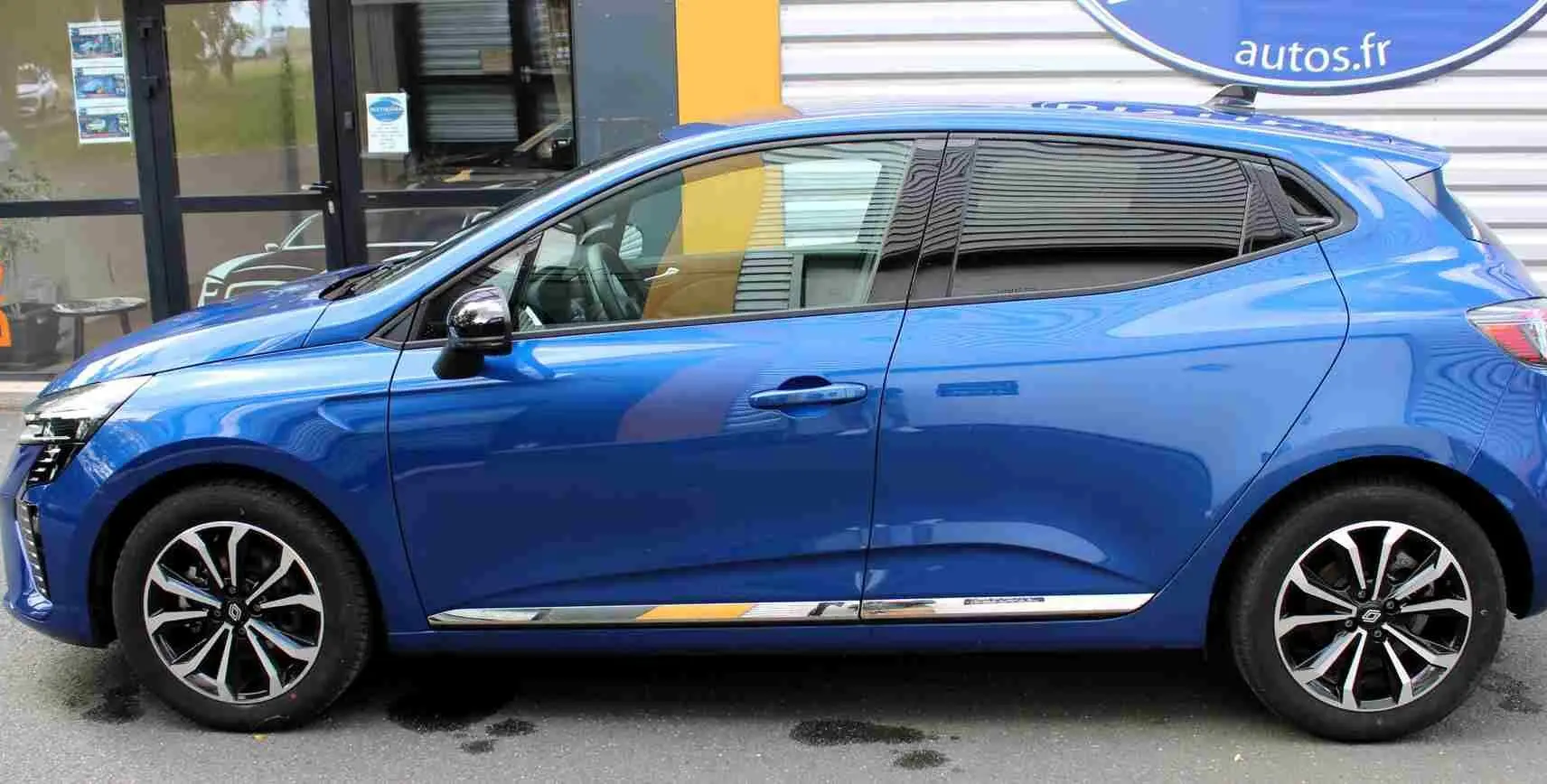 Profil droit de la Renault Clio 2025 bleu Iron Metal avec jantes alliage et vitres teintées, garée devant un bâtiment.