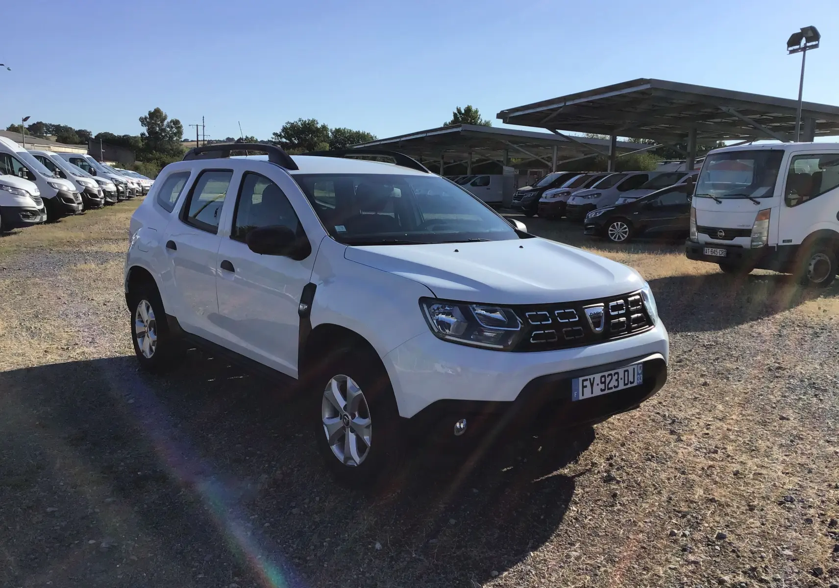 Dacia Duster blanc vu en 3/4 avant droit sur un terrain avec plusieurs véhicules en arrière-plan.