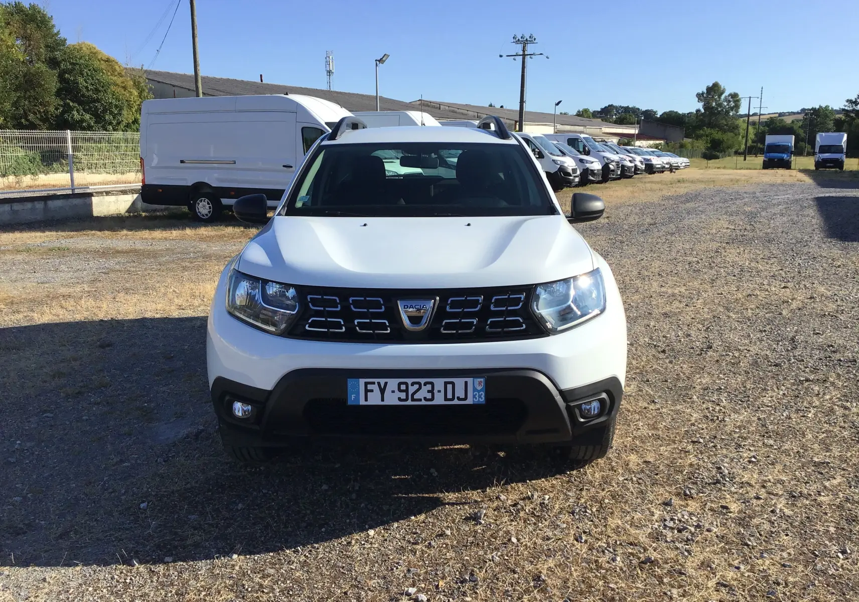 Vue de face d'un Dacia Duster blanc 2021 avec calandre noire et plaques d'immatriculation françaises sur un terrain extérieur.