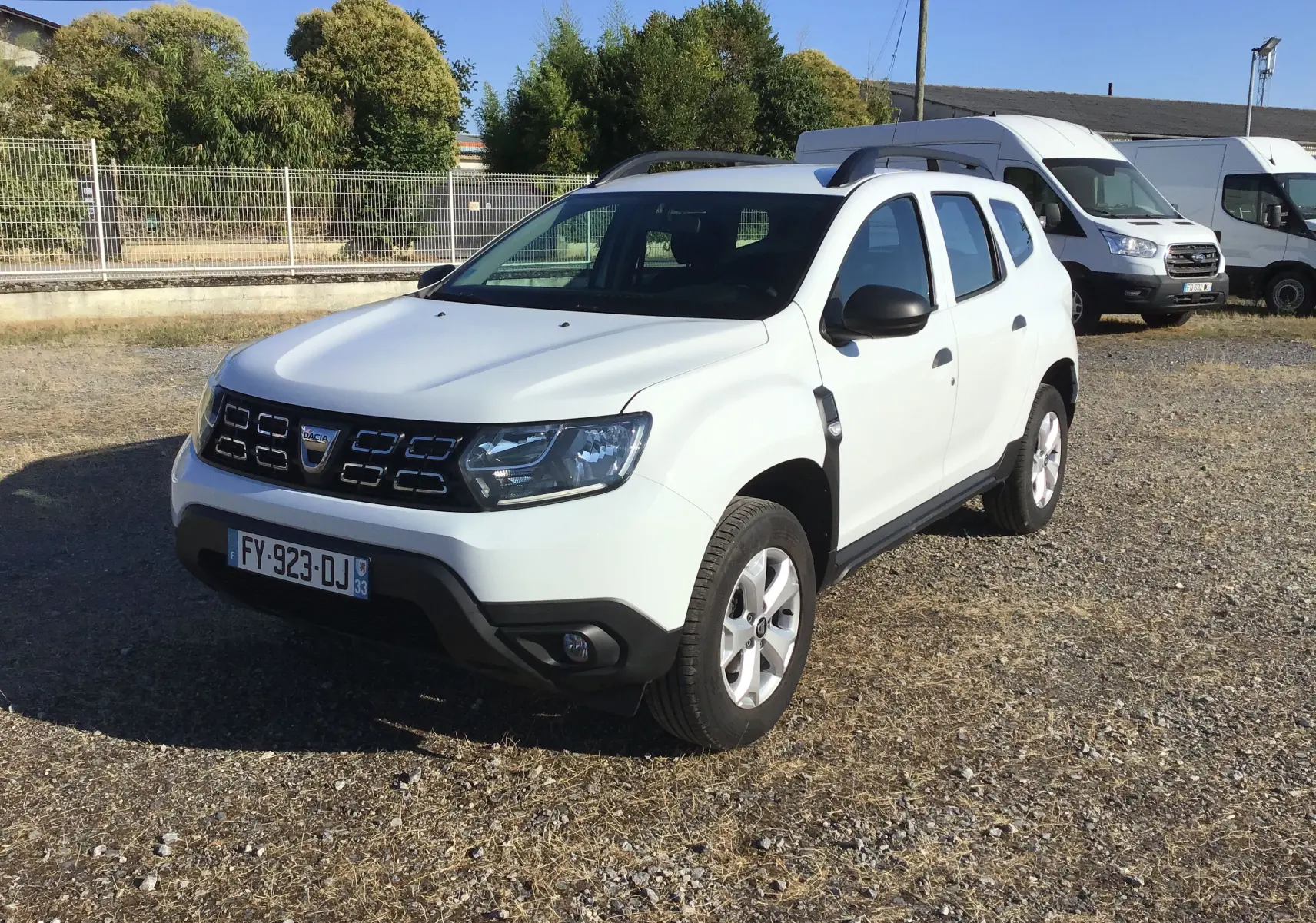 Dacia Duster blanc en 3/4 avant droit sur un sol sec, avec calandre noire et barres de toit visibles.