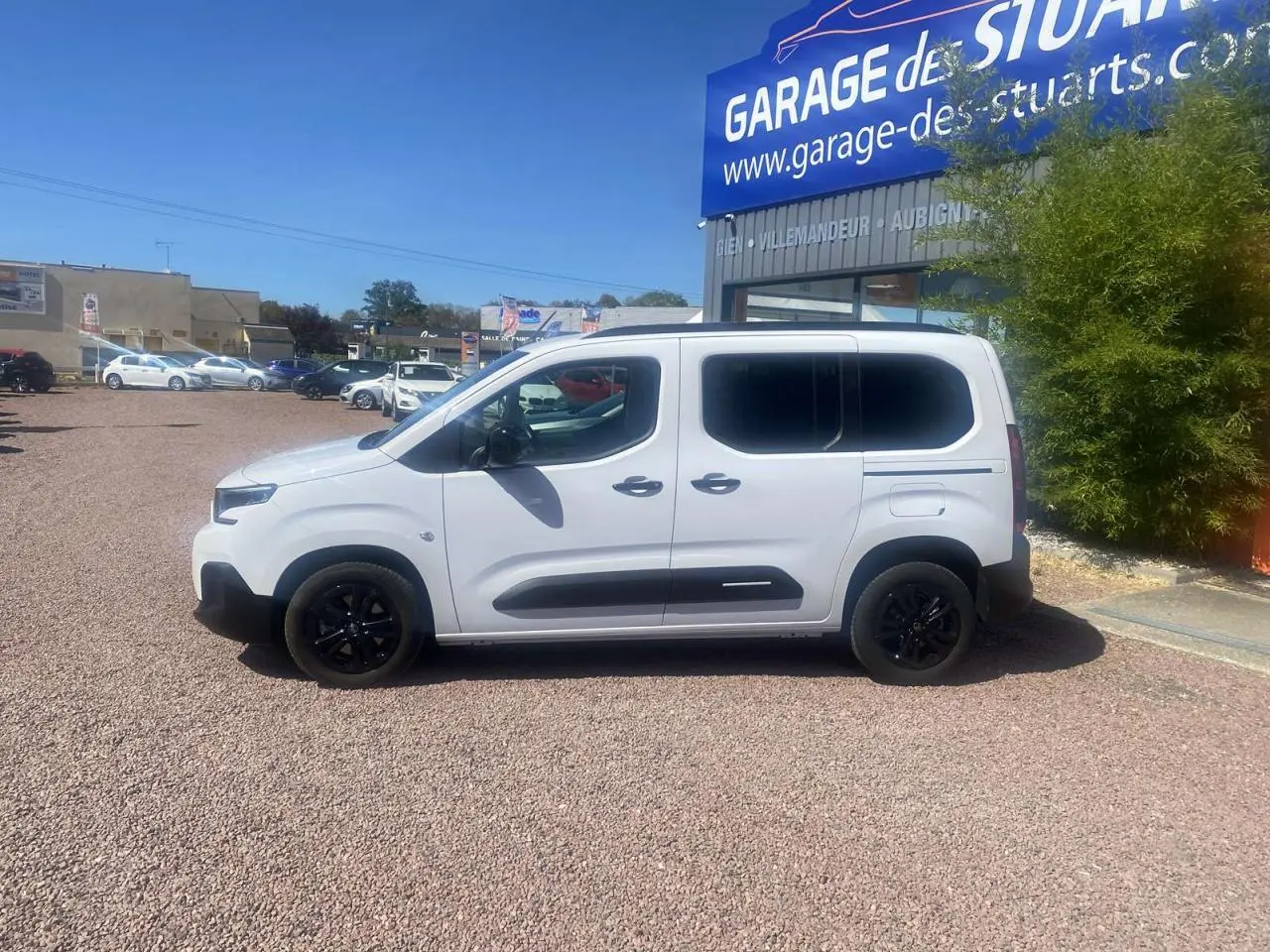Profil droit d'un Citroën Berlingo blanc 2025 avec portes coulissantes et jantes noires, stationné devant un garage.