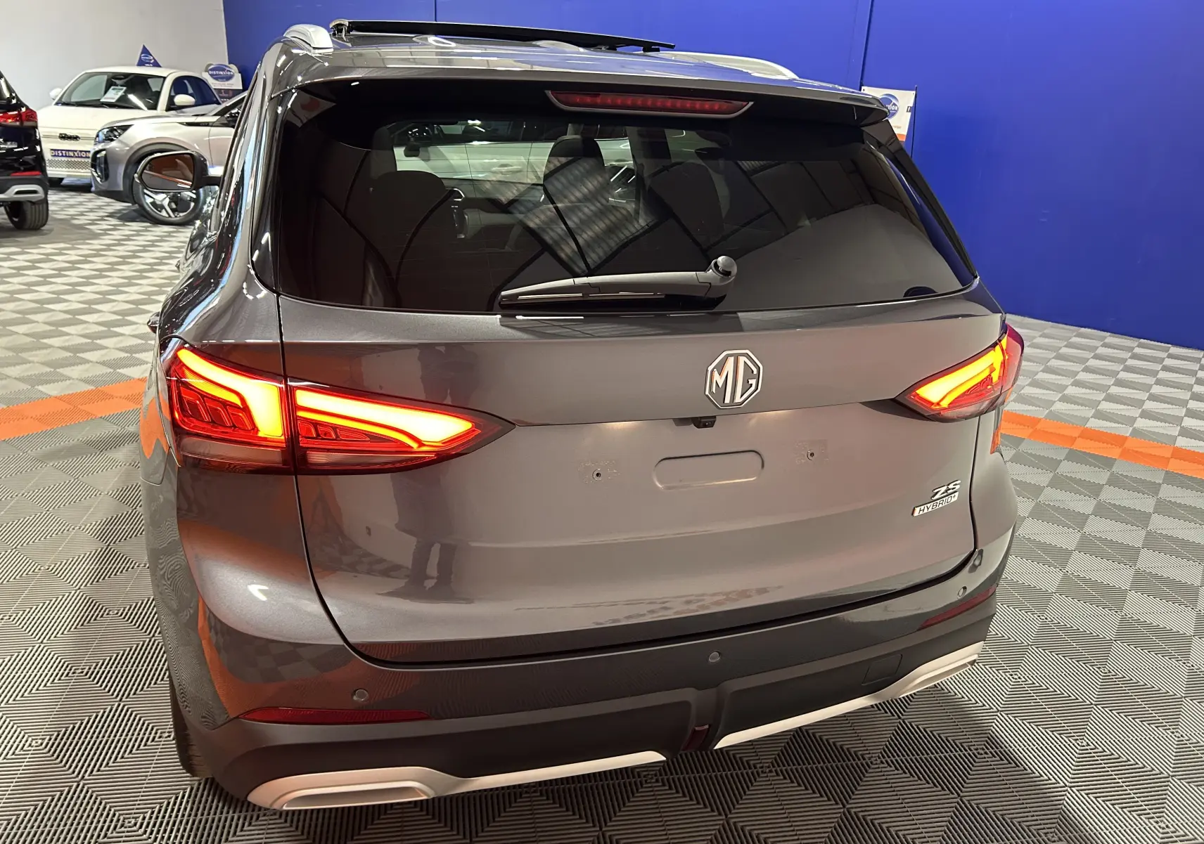 Vue arrière d'un MG ZS Hybrid+ 197 Luxury gris Candem avec feux allumés dans un showroom.