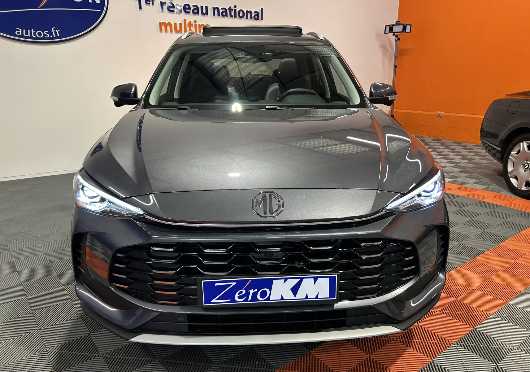 Vue avant du MG ZS Hybrid+ 197 Luxury gris Candem avec calandre noire et phares LED allumés dans un showroom.