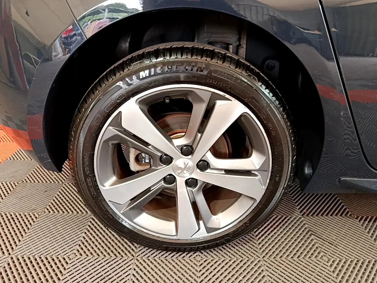Gros plan sur la roue avant gauche avec jante alliage argentée et logo Peugeot sur une 308 gris foncé.